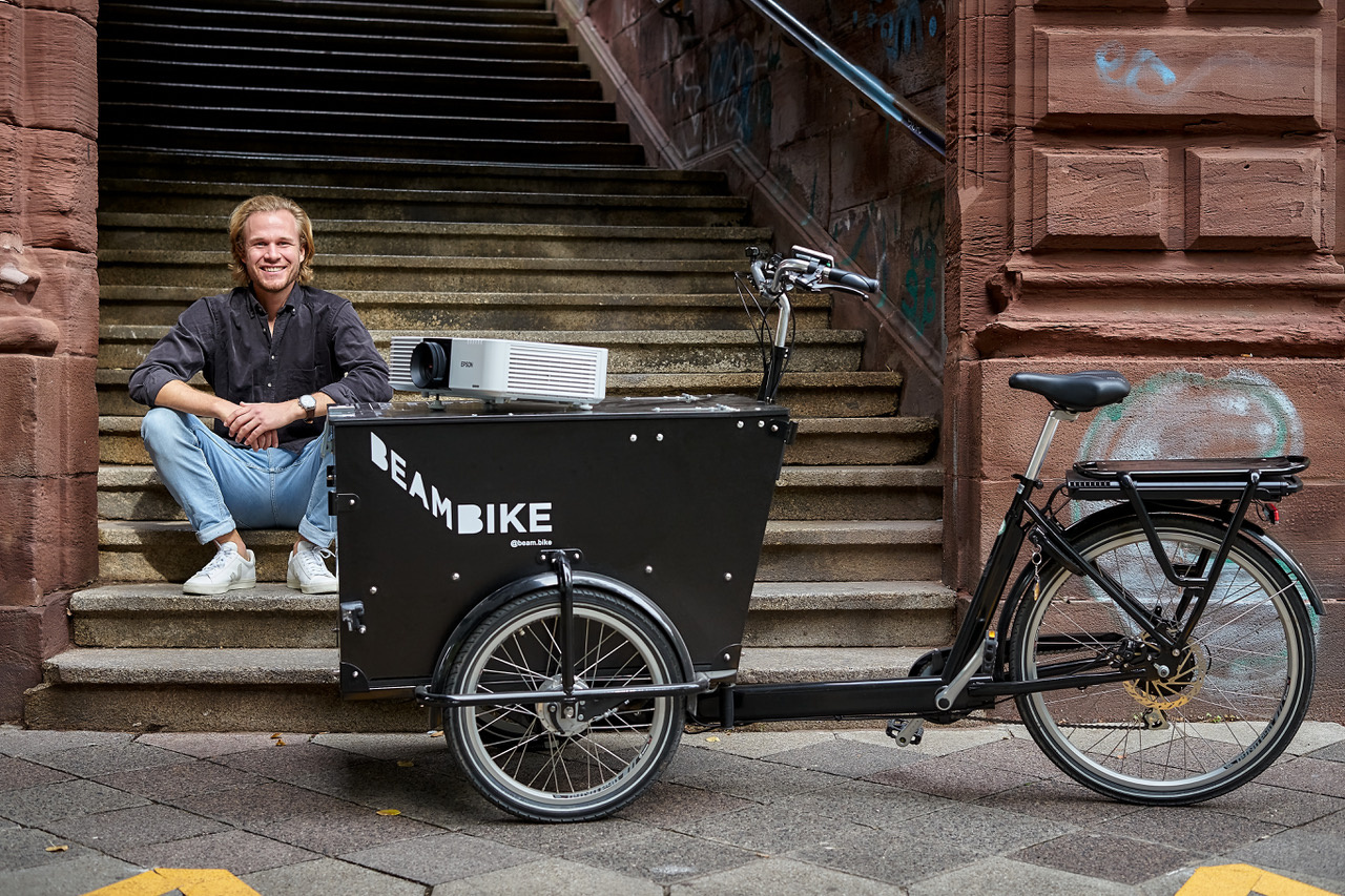 The photo shows a man sitting on a staircase. In front of him stands a bicycle with a beamer attached to it, the »beambike«.