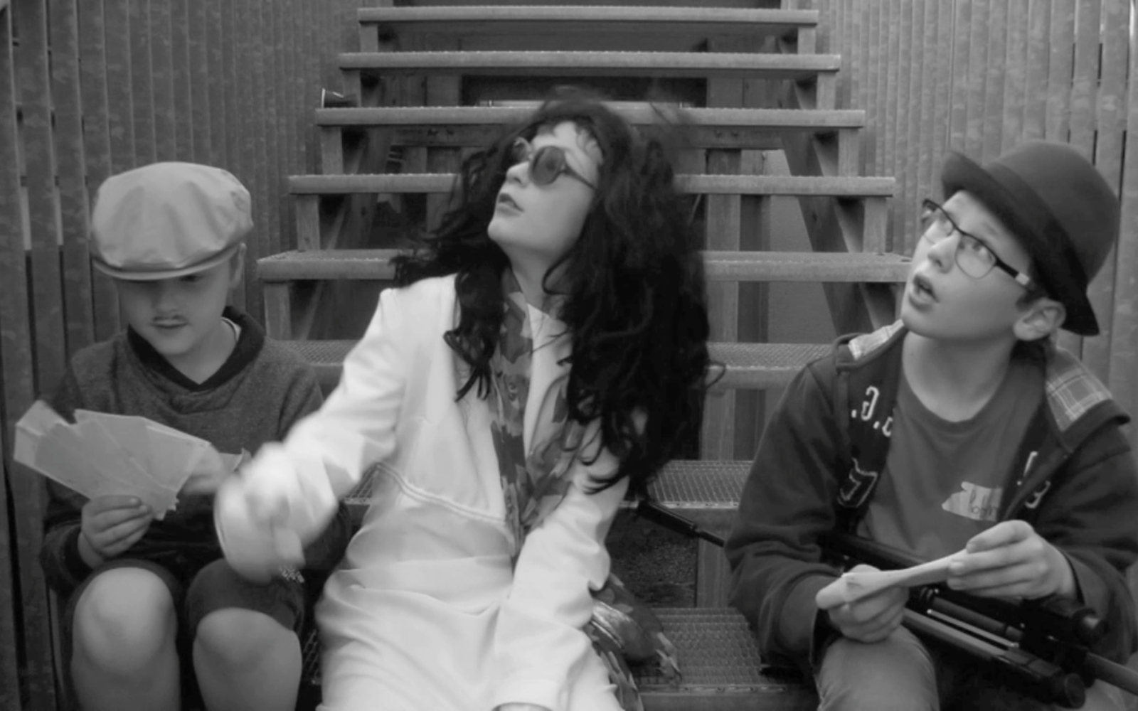 Three children are sitting in theatrical costumes on a staircase. Two are looking surprised upward and one is staring on fake money in his hands.