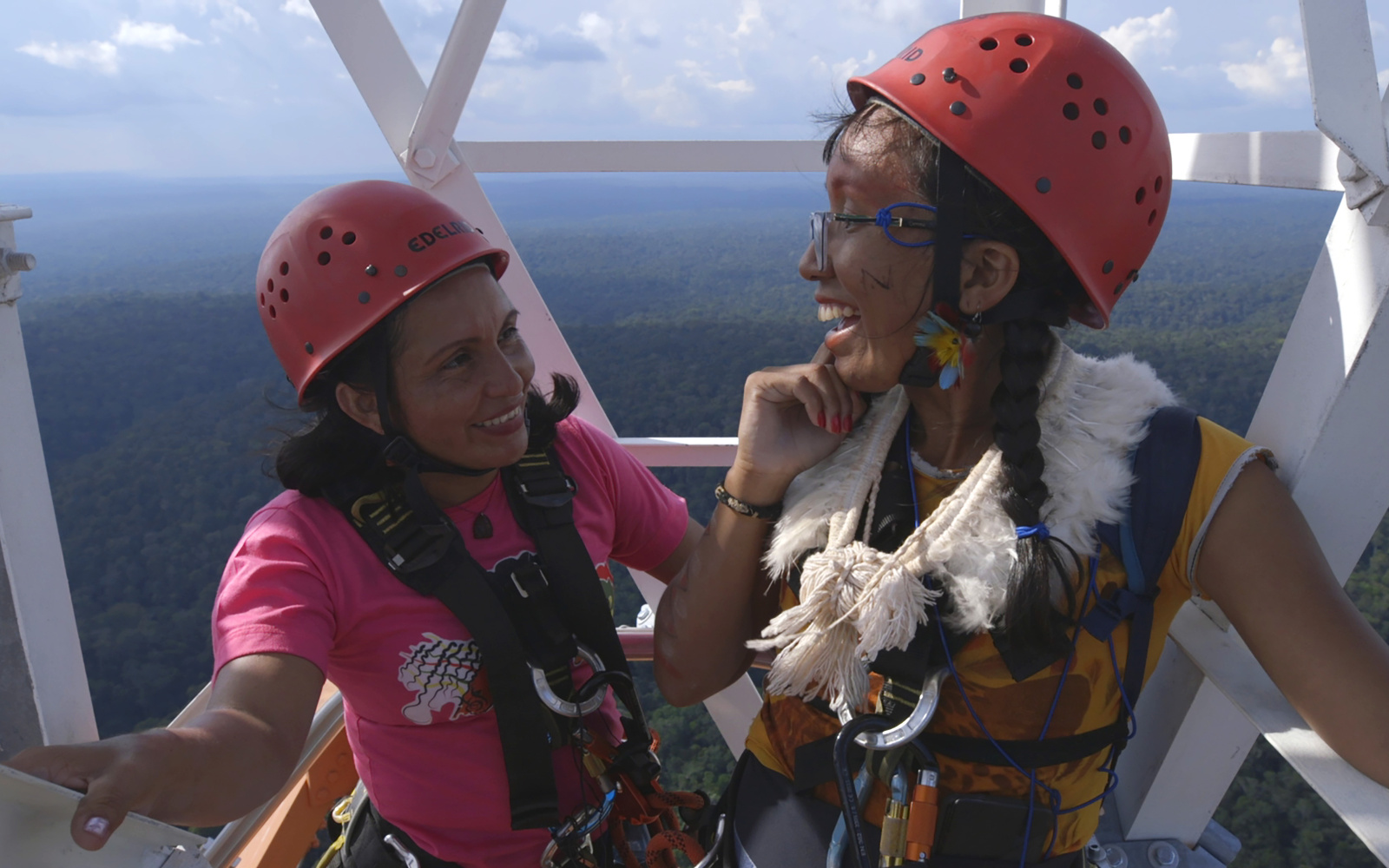 Two women can be seen standing high up on a tower. They wear safety helmets and laugh. In the background you can see forest up to the horizon.
