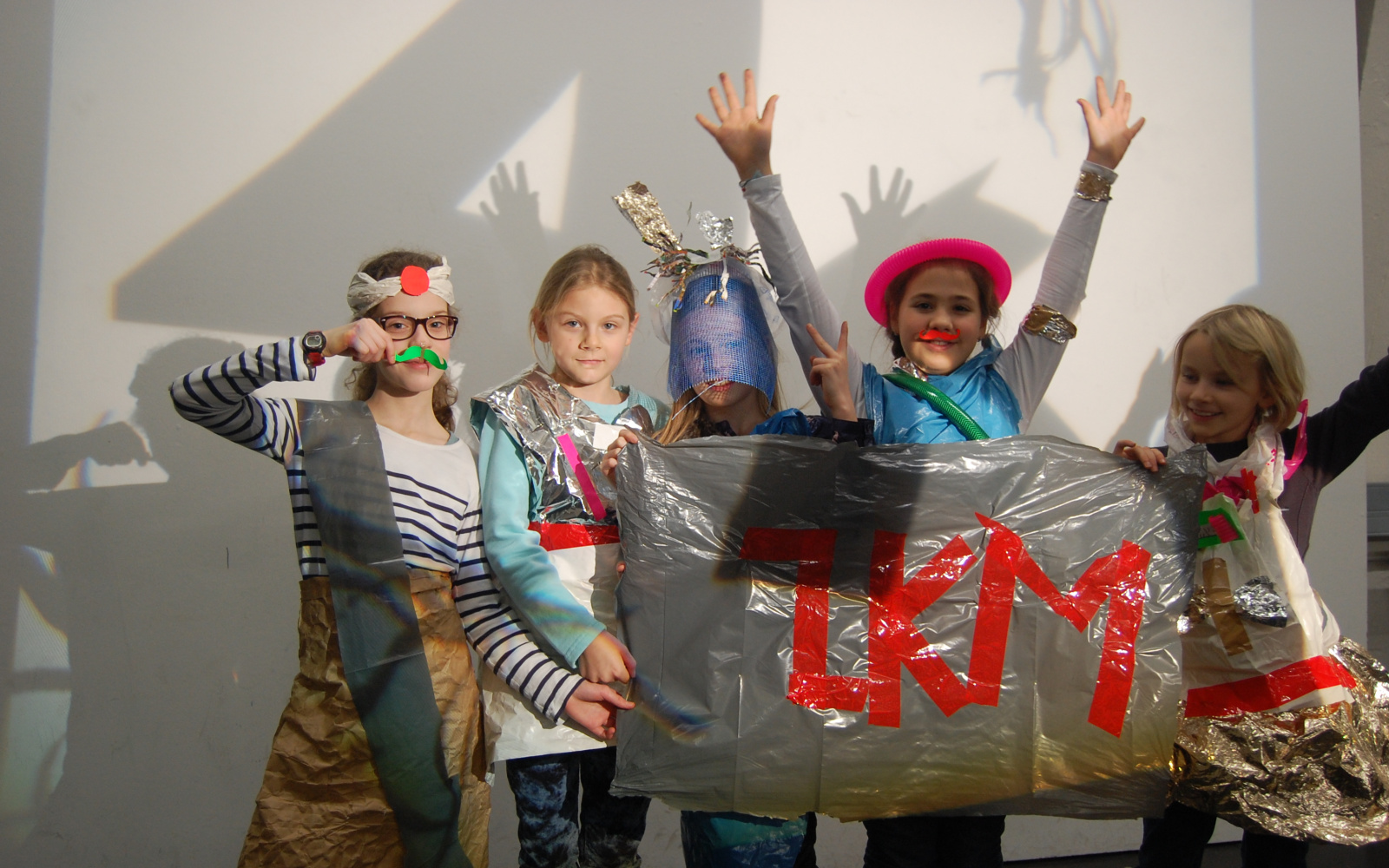 Eine Gruppe Mädchen, die sich aus alltäglichen Gegenständen Outfits gebastelt haben, halten ein Banner mit dem Schriftzug ZKM in rotem Klebeband vor sich.