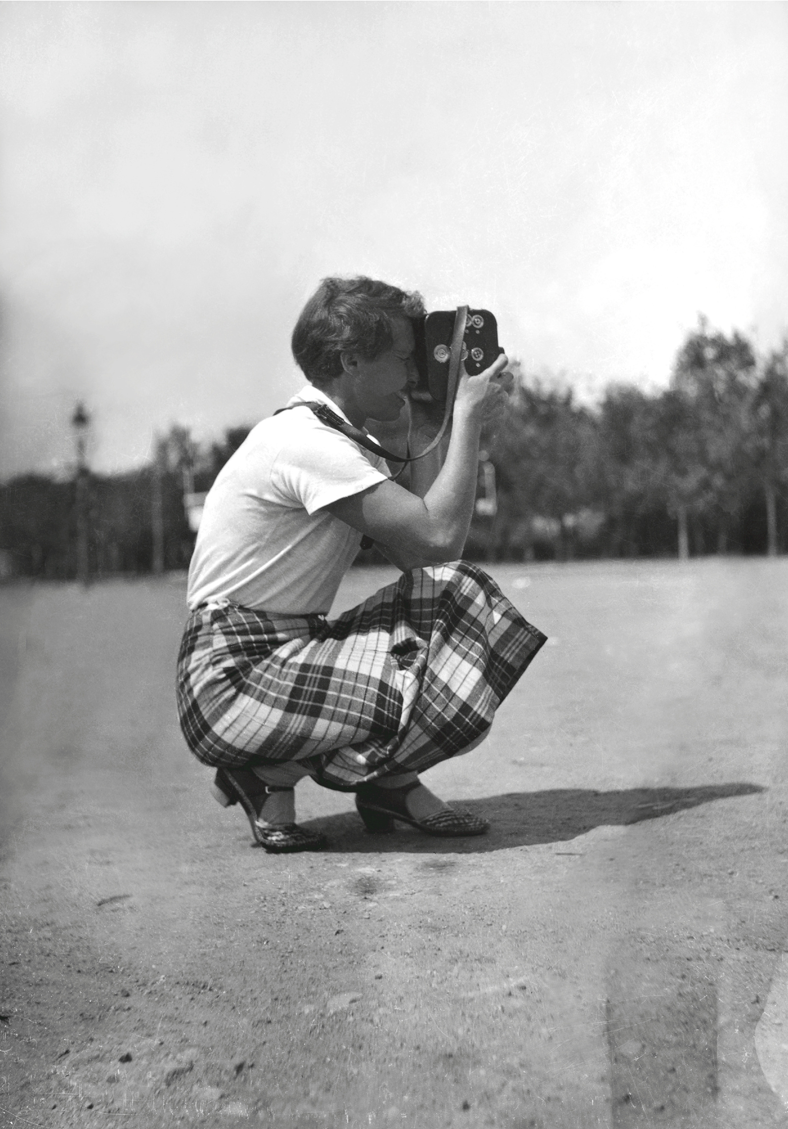 The black and white photograph shows a woman in squatting position with a camera.