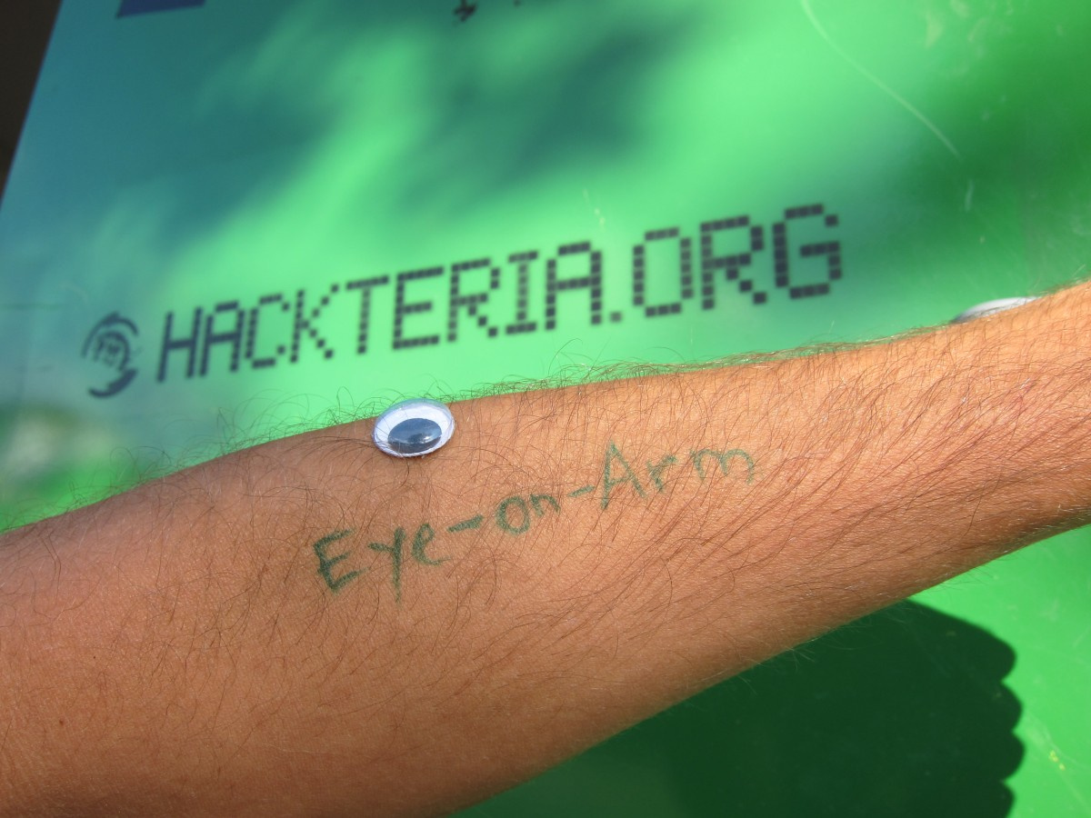 Auf einem Männerarm ist mit grüner Schrift Eye-on-Arm geschrieben und ein Spielzeugauge liegt darüber auf dem Unterarm. Der Hintergrund ist grün und mit der url hackteria.org beschrieben.