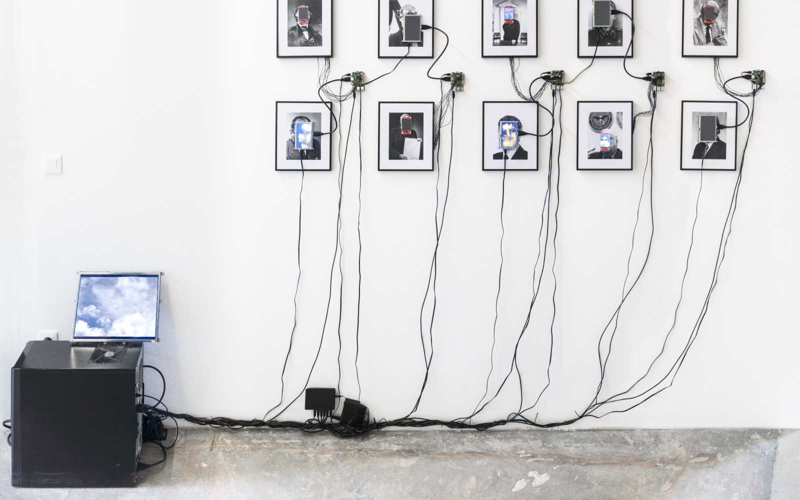 Das Foto zeigt 10 eingerahmte Portraits auf deren Gesichter kleine Monitore angebracht sind. Diese sind durch Kabel mit einem Computer verbunden.