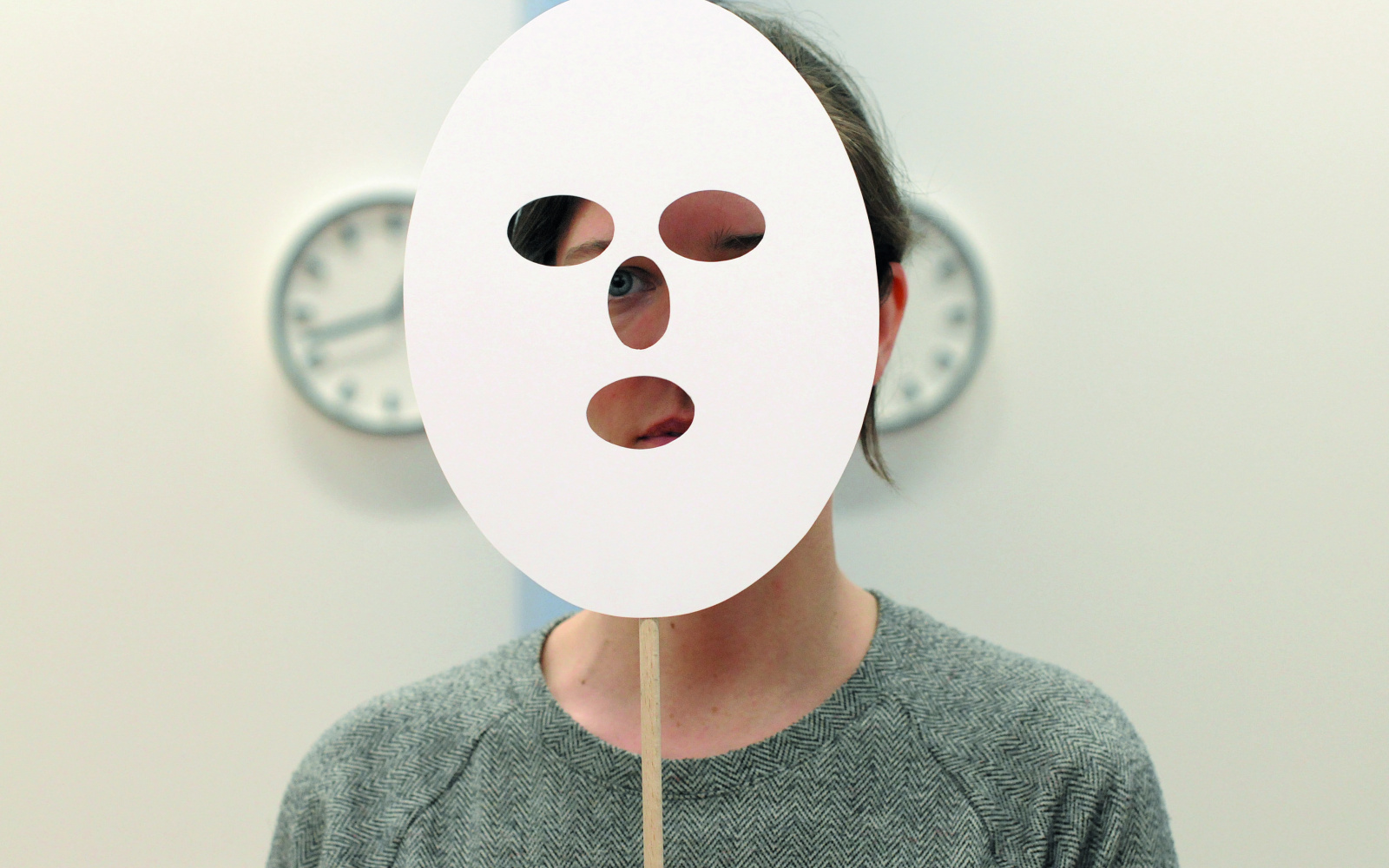 A person of undefined gender holding a white paper mask in fron of her face.