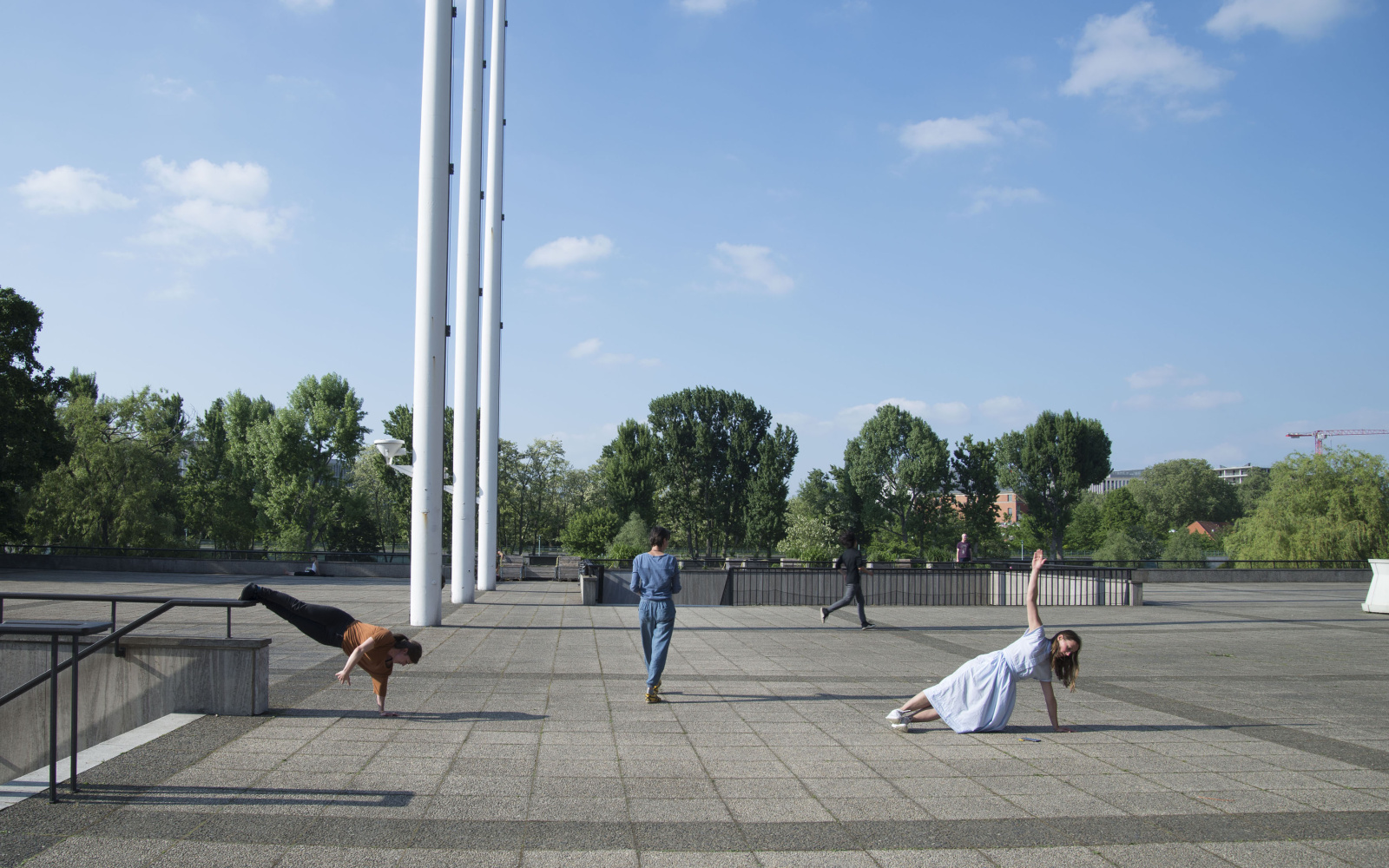 You can see a large, paved square in the open. In the background are some trees. In the picture 3 persons are standing far apart. The two people outside are doing a push-up, the person in the middle turns his back to the camera. 