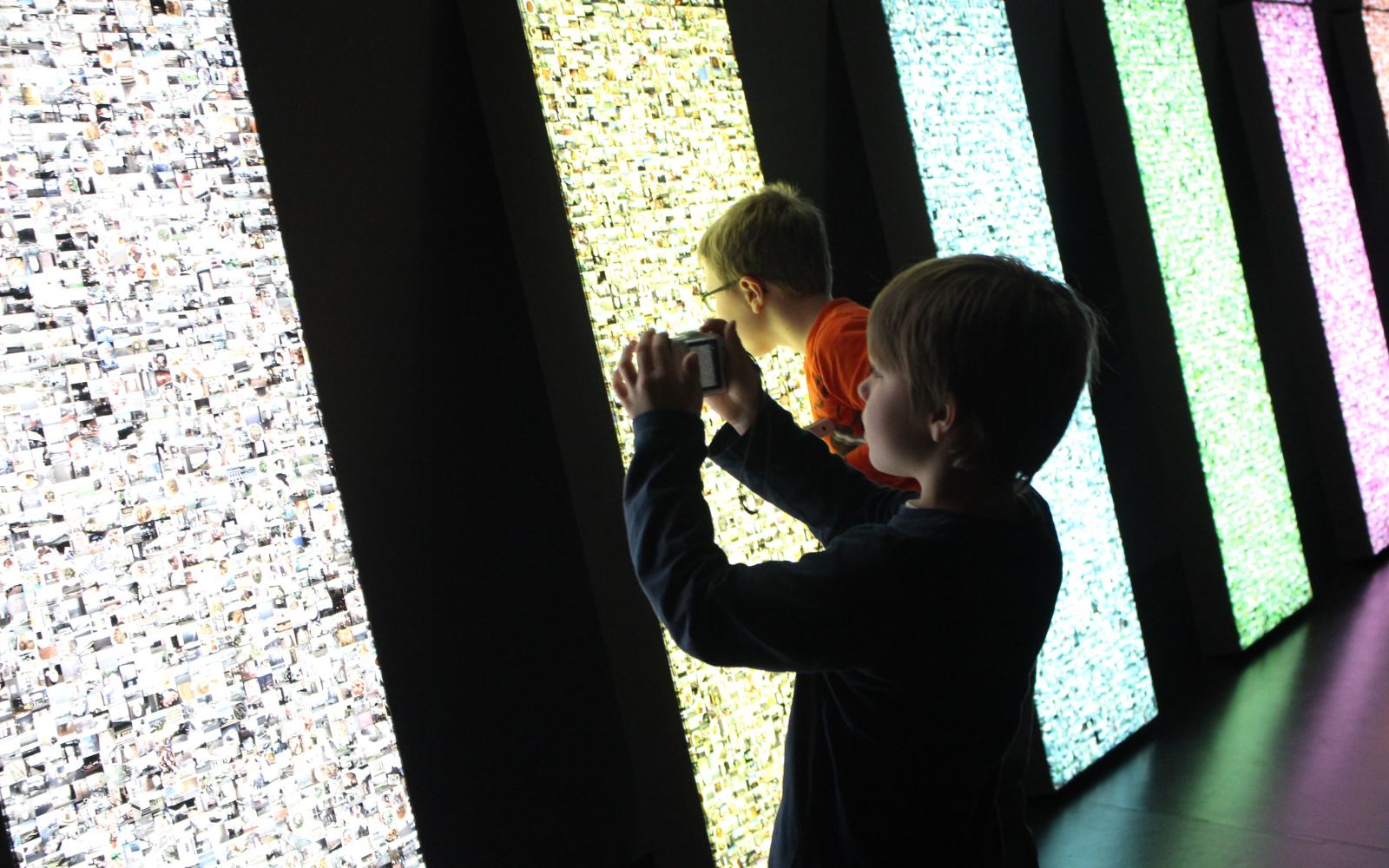 Two kids are standing in front of an illuminated wall. One of them is taking a photo of the installation and the other has his face very close to the work of art to look closely at the little pictures, that are beeing illuminated.