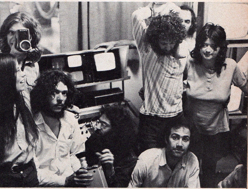 The black-and-white image shows a group of long-haired young people in front of small screens