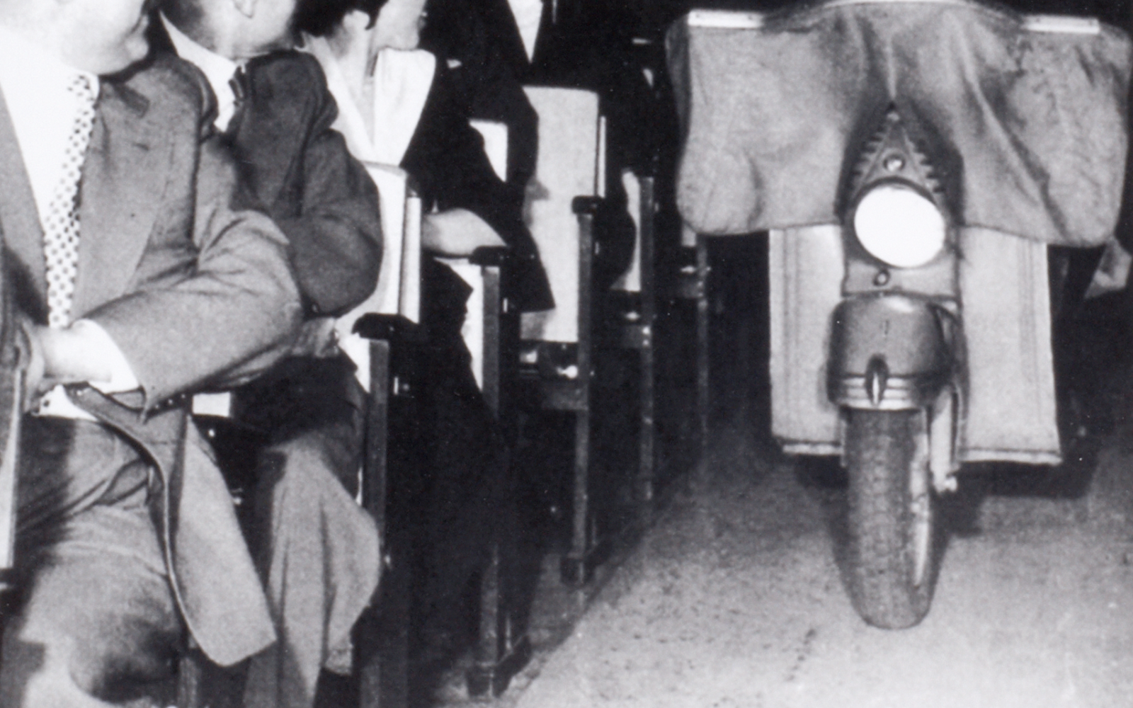  A black-and-white photo shows a man riding a moped into a room where many people sit on chairs.
