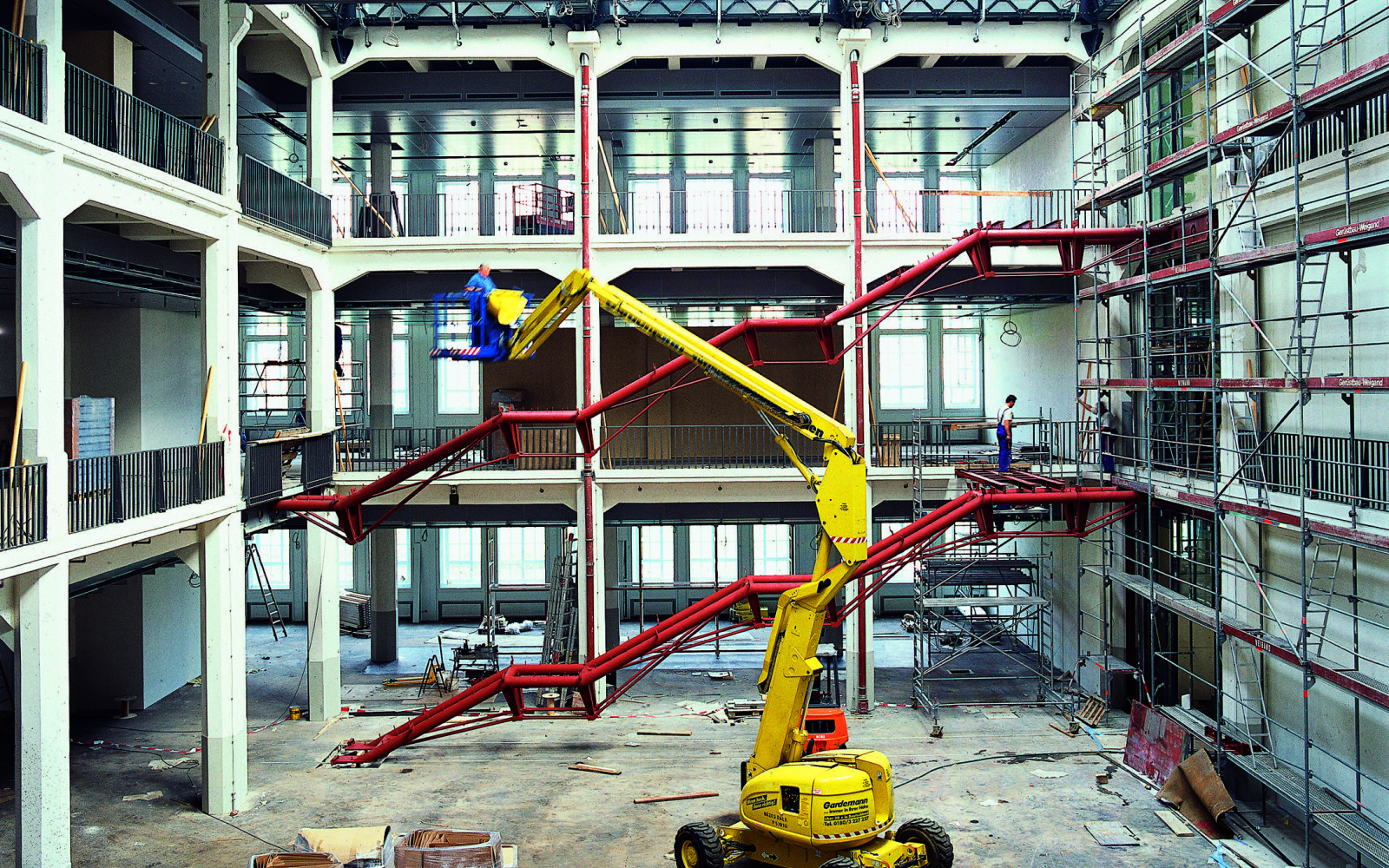 Das Foto zeigt eine historische Ansicht des Umbaus des alten MNK Hallenbaus. Ein imposanter großer gelber Bagger steht mitten in der Halle während im Hintergrund die Rohkonstruktion der zukünftigen Treppen des Ausstellungsraumes in rot hell leuchten.