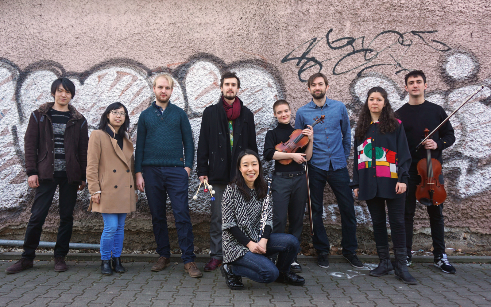Das Bild zeigt die StipendiatInnen der Internationalen Ensemble Modern Akademie (IEMA) vor einer vollgesprühten Backsteinwand