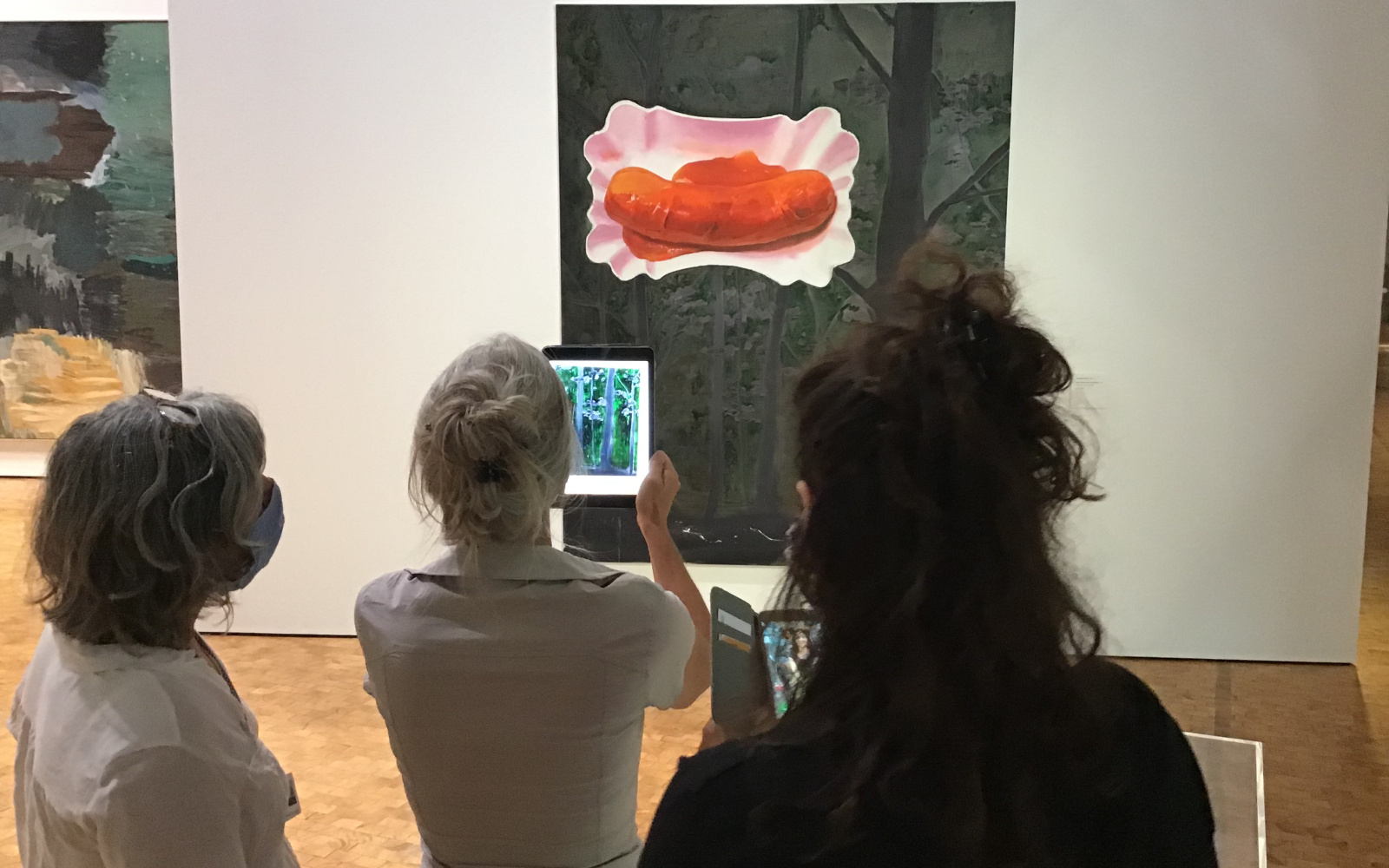Three women stand in front of a picture showing a curry sausage in a cardboard bowl. One of the women holds an iPad on the picture.