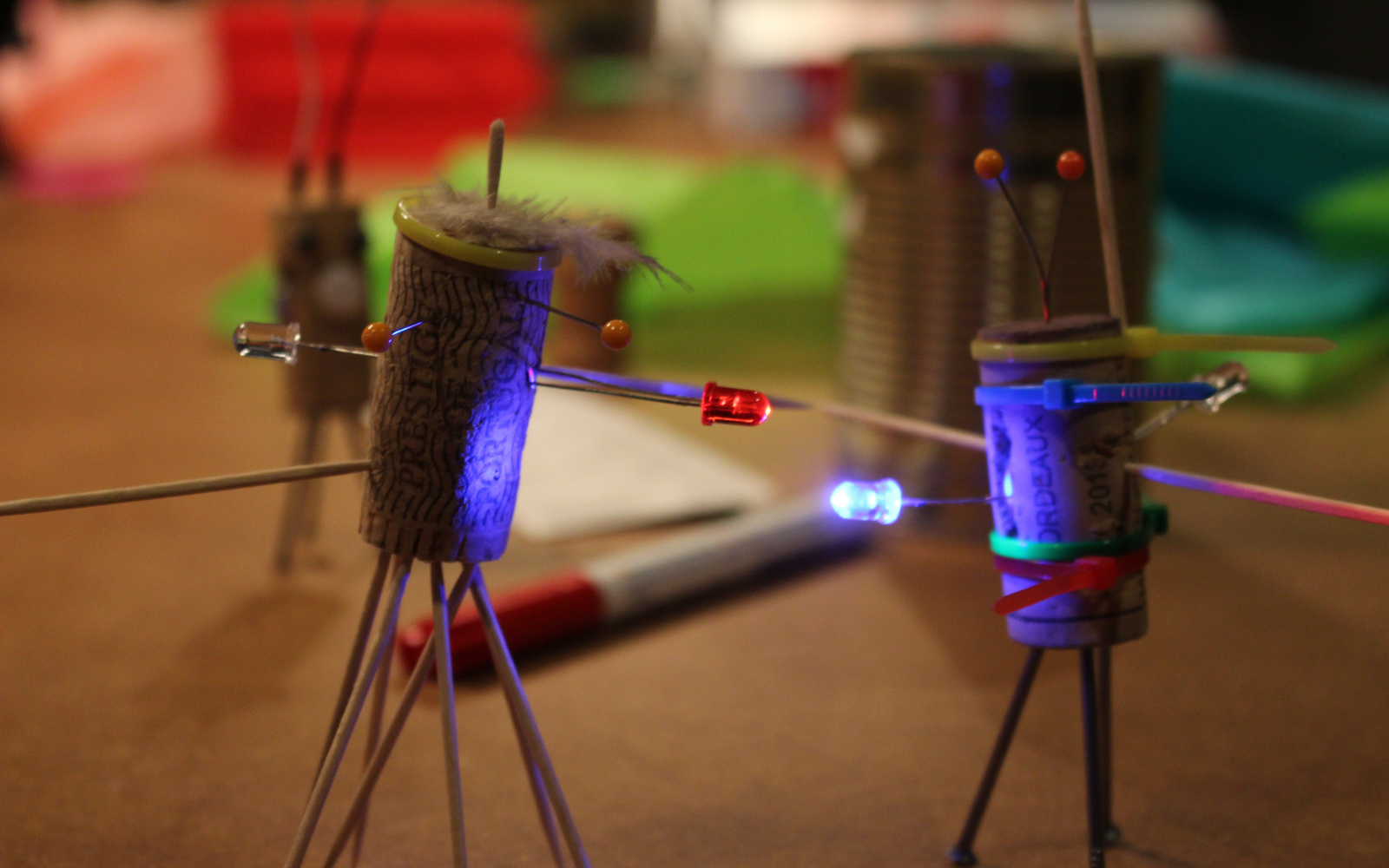 Zwei Korken, die zu kleinen Figuren umgewandelt wurden und mit LEDs ausgestattet sind stehen auf einem Tisch mit allerlei Bastelutensilien.