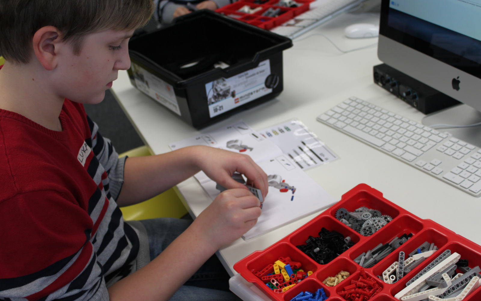 A boy is constructing a lego-robot. On the table he is sitting at are boxes filled with robot parts.