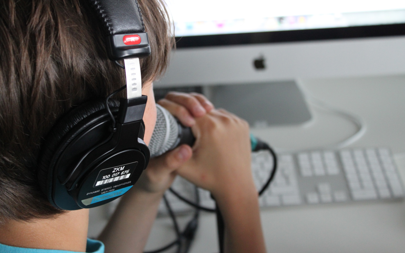 Ein Junge spricht in ein Mikrofon, das er mit beiden Händen vor den Mund hält. Er schaut dabei auf den Computerbildschirm vor ihm.