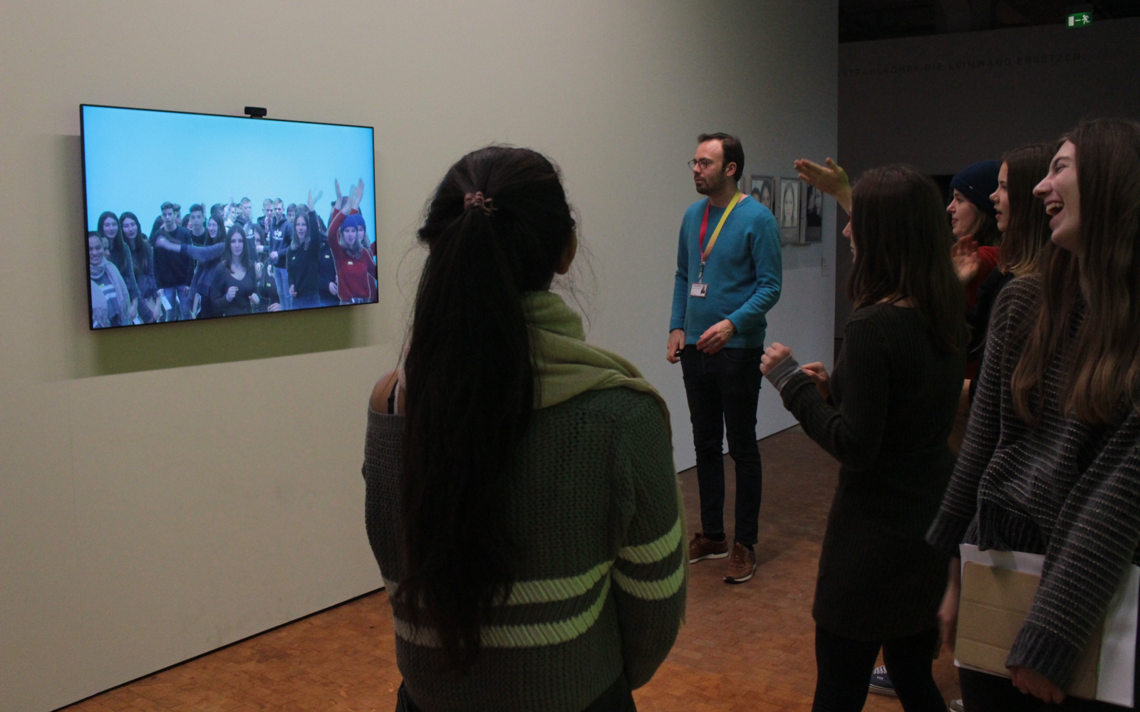 Zu sehen ist eine Gruppe an jungen Menschen, die vor einem Bildschirm stehen. Auf dem Bildschirm ist ein Live-Bild von der Gruppe zu sehen, das von einer Kamera über dem Bildschirm aufgenommen wird.