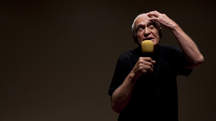 A man speaks into a microphone and holds up his left hand to his head