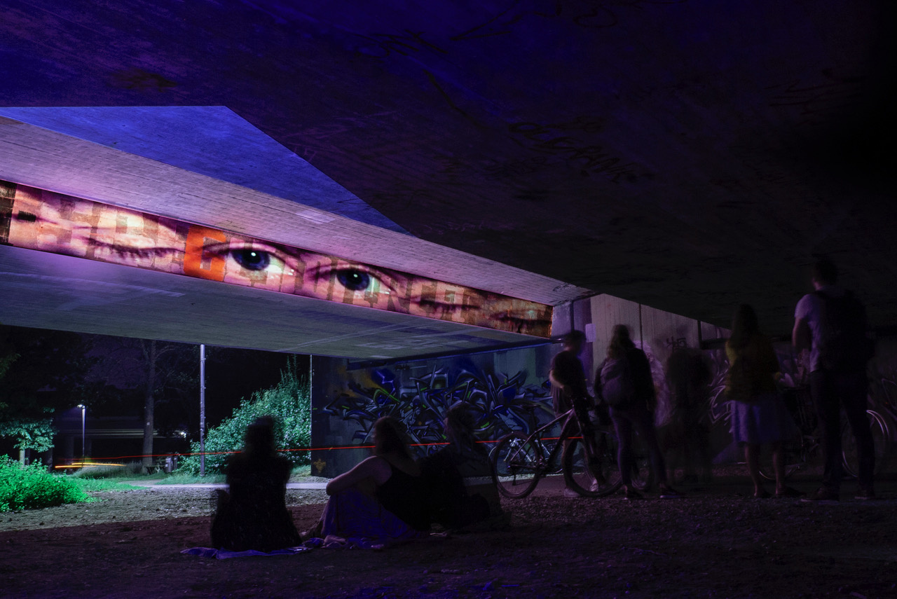 Eine Brücke in einem Park bei Nacht. An die Außenwand der Brücke ist in einem horizontalen Streifen eine Reihe Augenpaare projiziert. Der Rets der Brücke leuchtet in verschiedenen Farben.