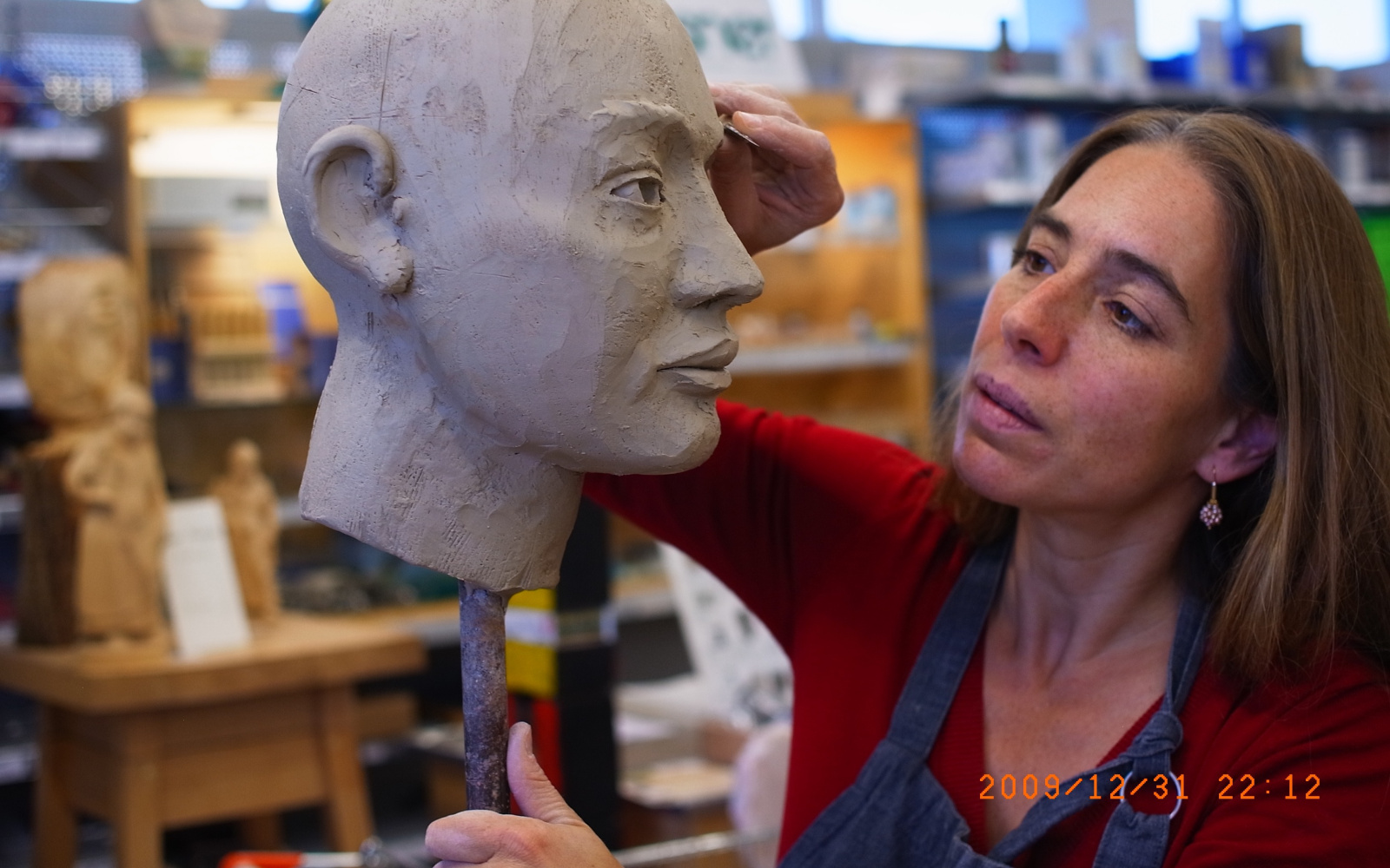 A model of a head sticking on a pole. A woman is working on it with a sculpting tool.