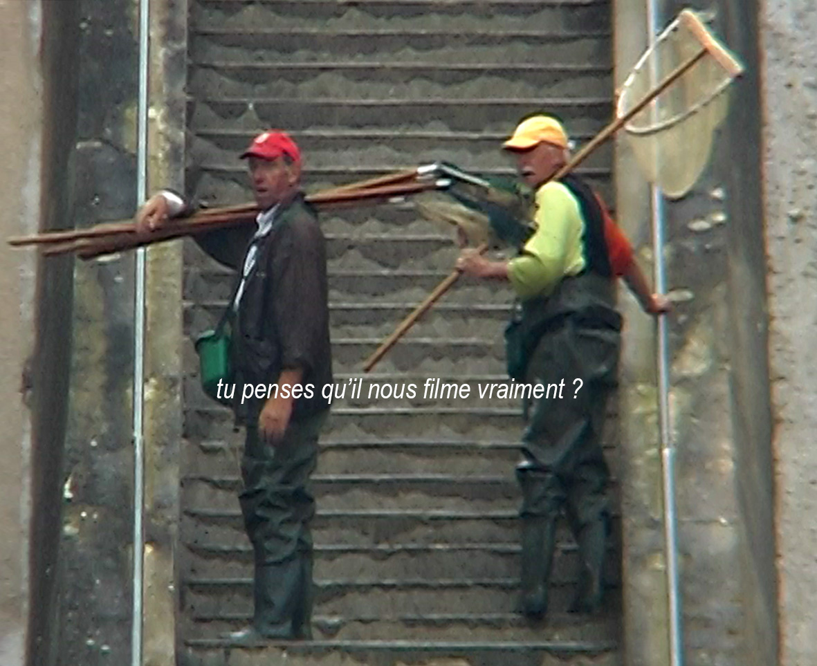 A film still from the film Héros absurde by Edmund Kuppel shows two men at the foot of an outdoor stone staircase, one on the left, the other on the right. They wear caps and fishing gear. They ask: "Tu penses qu'il nous filment vraiment?"