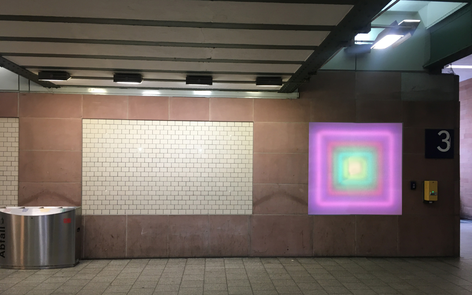 Zu sehen ist der Durchgang unter dem Karlsruher Hauptbahnhof. An der Wand ist ein leuchtendes Quadrat angebracht, das von innen nach außen einen Farbverlauf hat, der eine Tiefe erzeugt.
