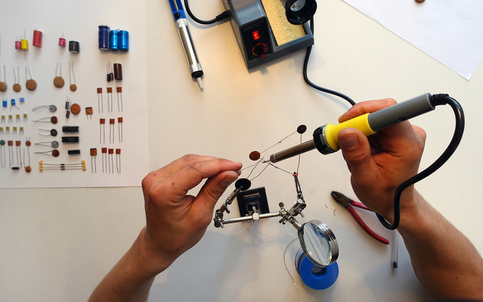 You can see hands from above that are holding a soldering iron. The person is soldering some wires together using a estra hand..