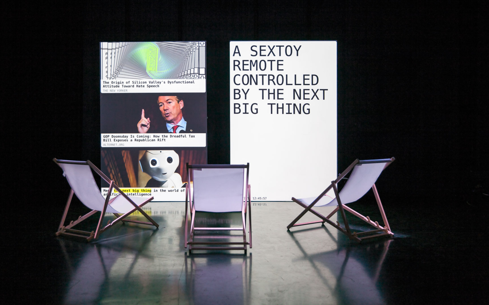 The photo shows three deck chairs in front of two upright rectangular canvases. On the left screen you can see a spiral form, the head of a man and the head of a robot. A text appears on the right screen. 