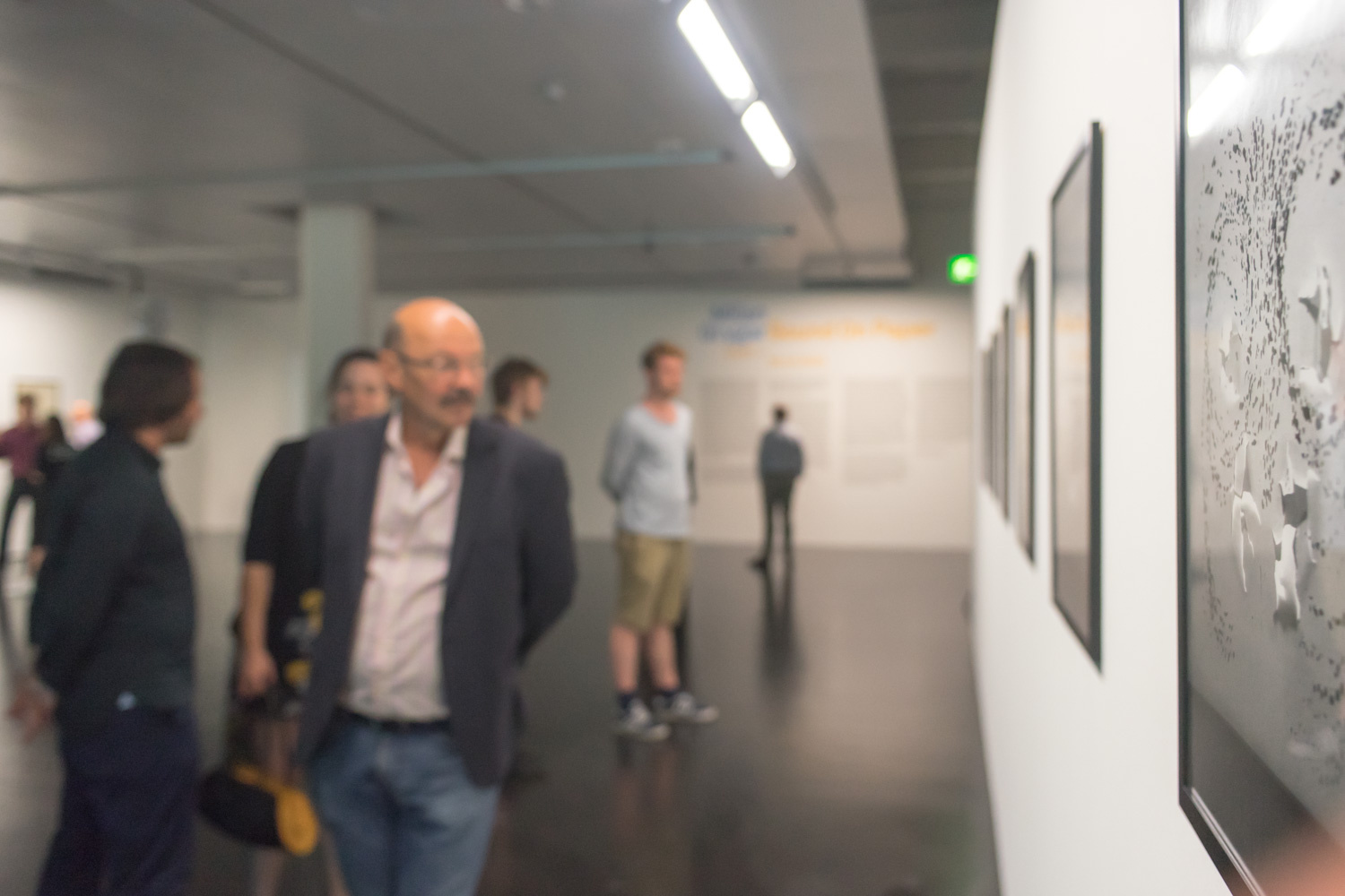 People in the museum at the Vernissage are blurry in the picture, but the artwork on the wall is sharp and shows abstract forms in shades of gray.