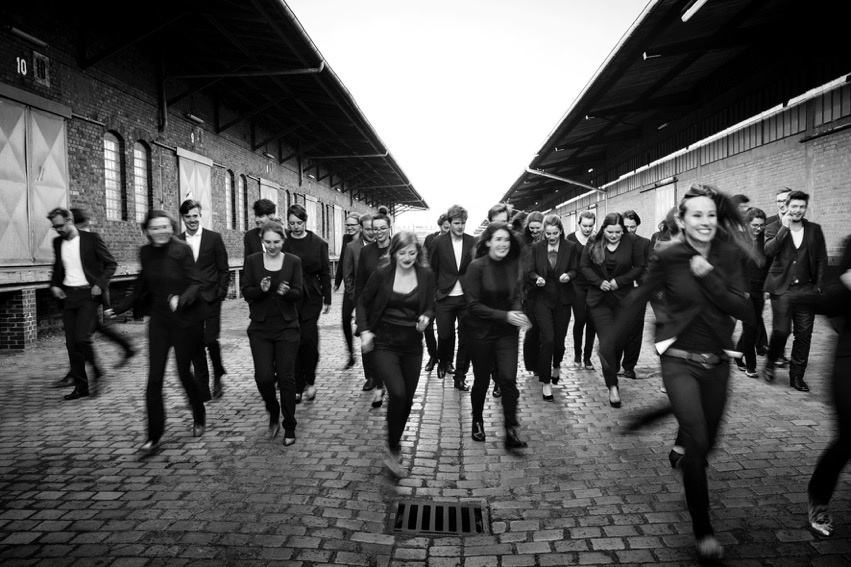 The black-and-white picture shows the members of the Ensemble Reflektor running across a paved street. 