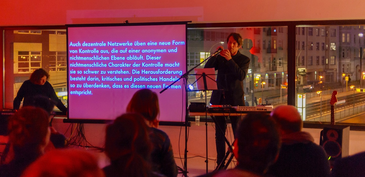Ein Mann bei seiner Performance am Keyboard und hinter ihm eine Leinwand, auf die ein Text projiziert ist.