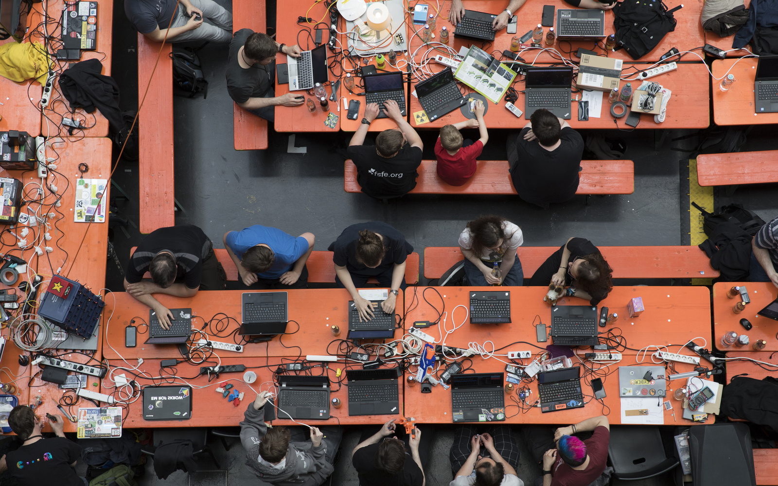 A bird's eye view of various orange beer benches where different people sit and operate different computers.