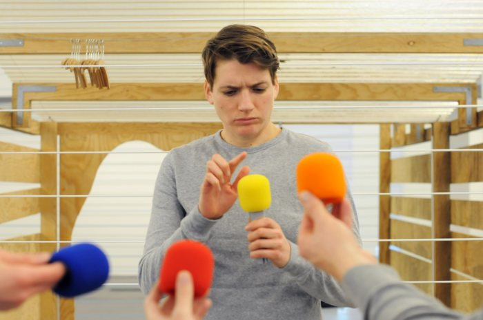 Ein junger Mann tippt kritisch mit seinem Finger auf ein gelbes Mikrofon. Er ist von verschiedenen Mikrofonen umgeben.