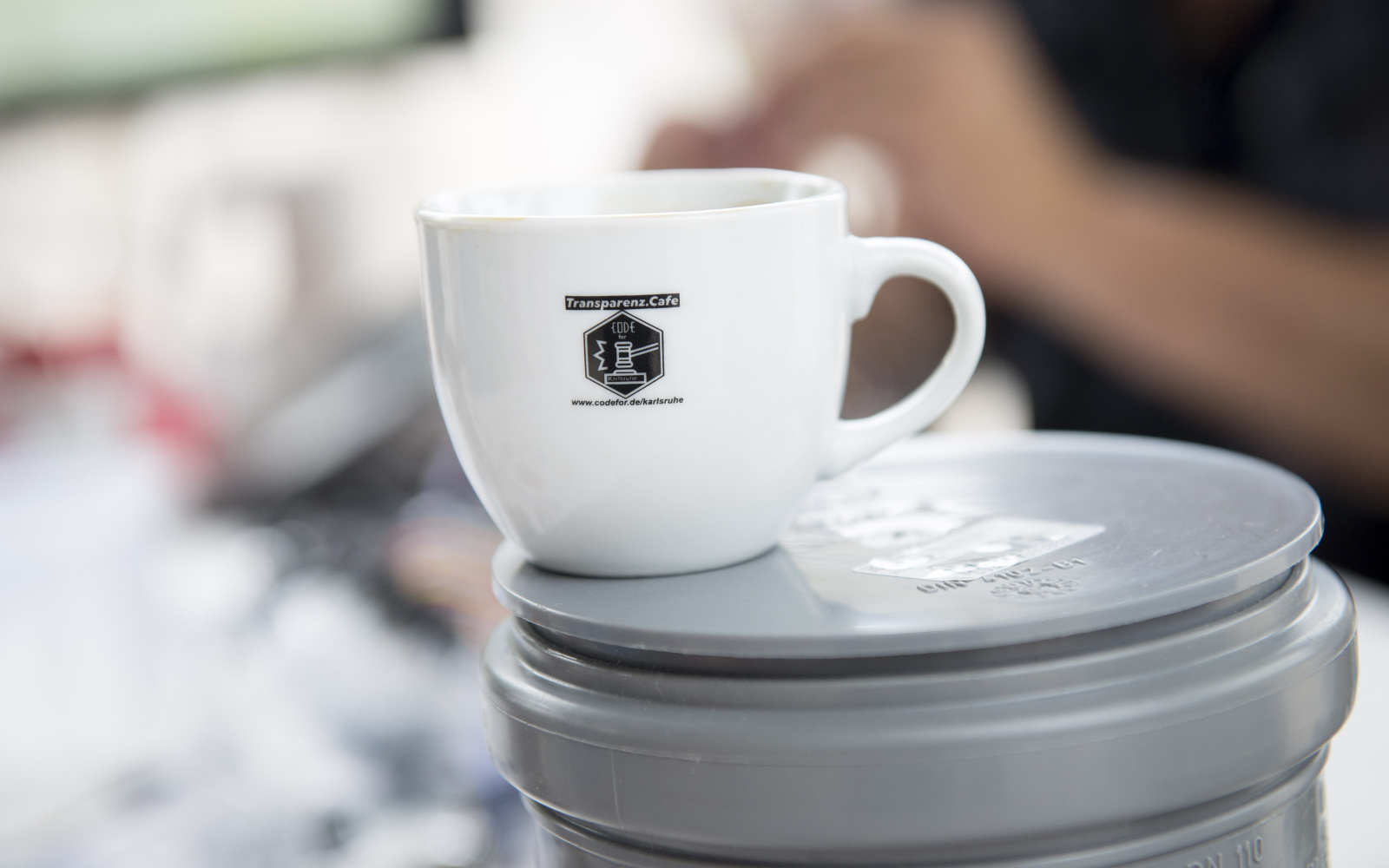 Zu sehen ist eine weiße Kaffetasse auf der das Transparenz Cafè Logo zu sehen ist.