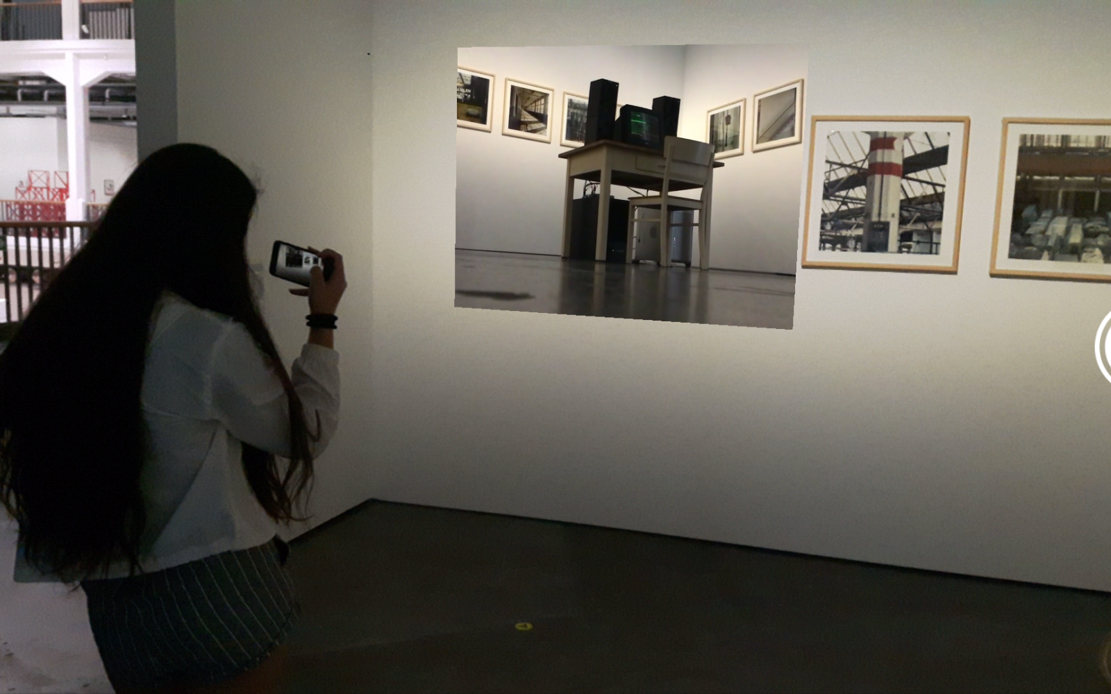 A girl is standing in front of a picture with her cell phone in her hand. She holds her cell phone as if she was filming the picture.