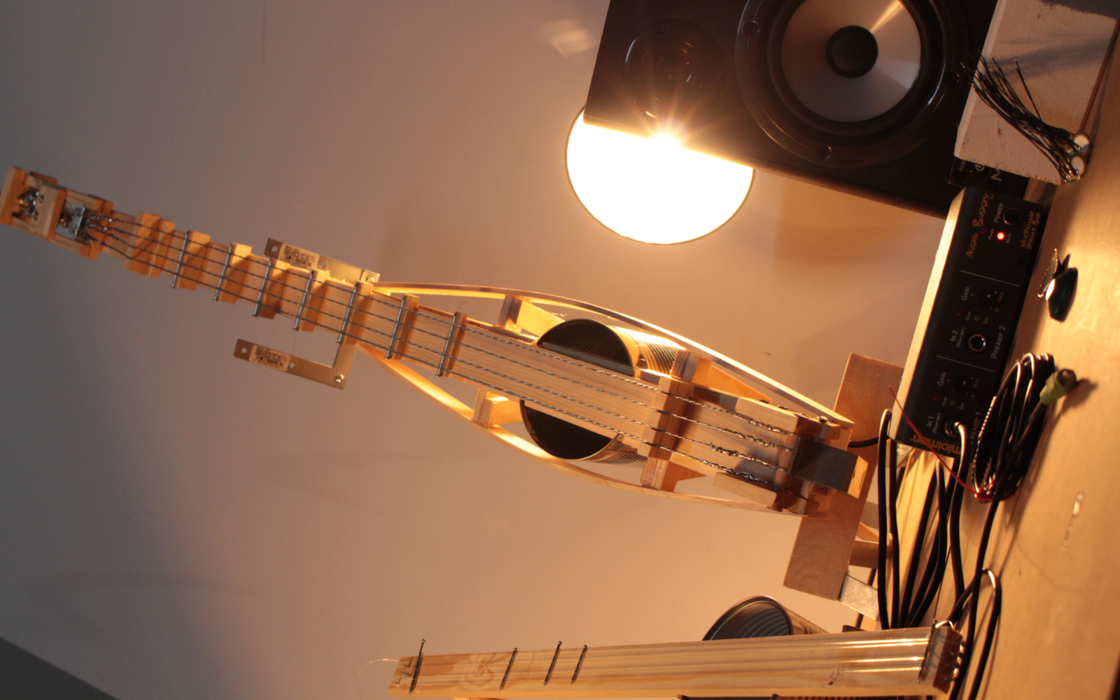 Eine aus dünnen Holzplatten und einer Konservendose bestehende selbst gebastelte Gitarre wird von einer Lampe angestrahlt. Sie steht aufrecht und daneben ist ein Lautsprecher aufgestellt.