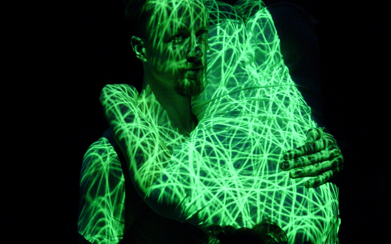 Kenneth Flak stands upright, in his left arm he holds Külli Roosna, she hugs him. Both are covered by a projection with neon green lines.