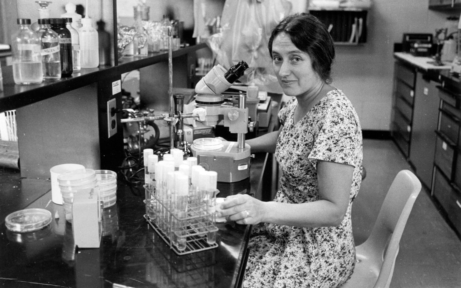 Die junge Lynn Margulis sitzt in einem Labor mit zahlreichen Laborutensilien. Sie trägt ein Sommerkleid und lächelt leicht.