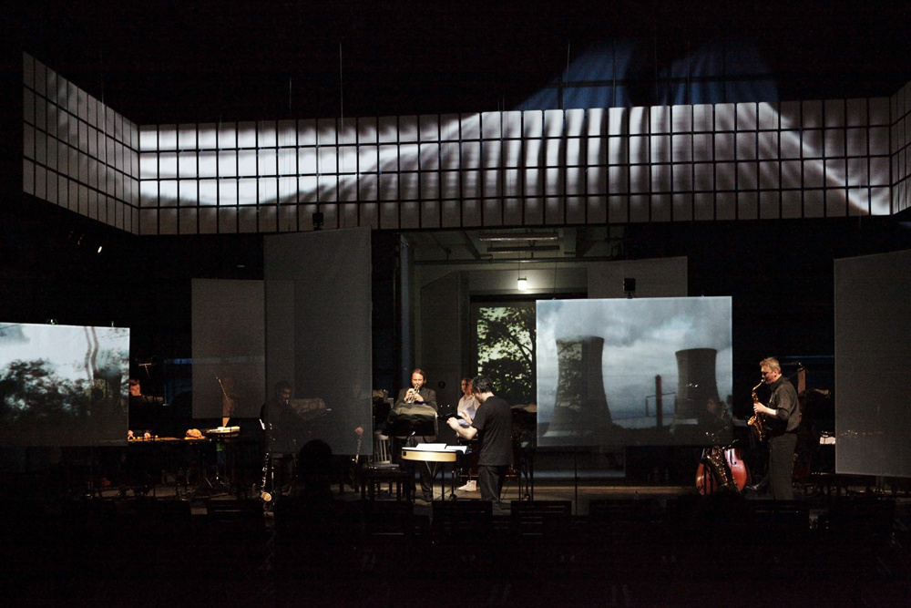 A music ensemble on a dusky stage. In the background a picture of a nuclear power station