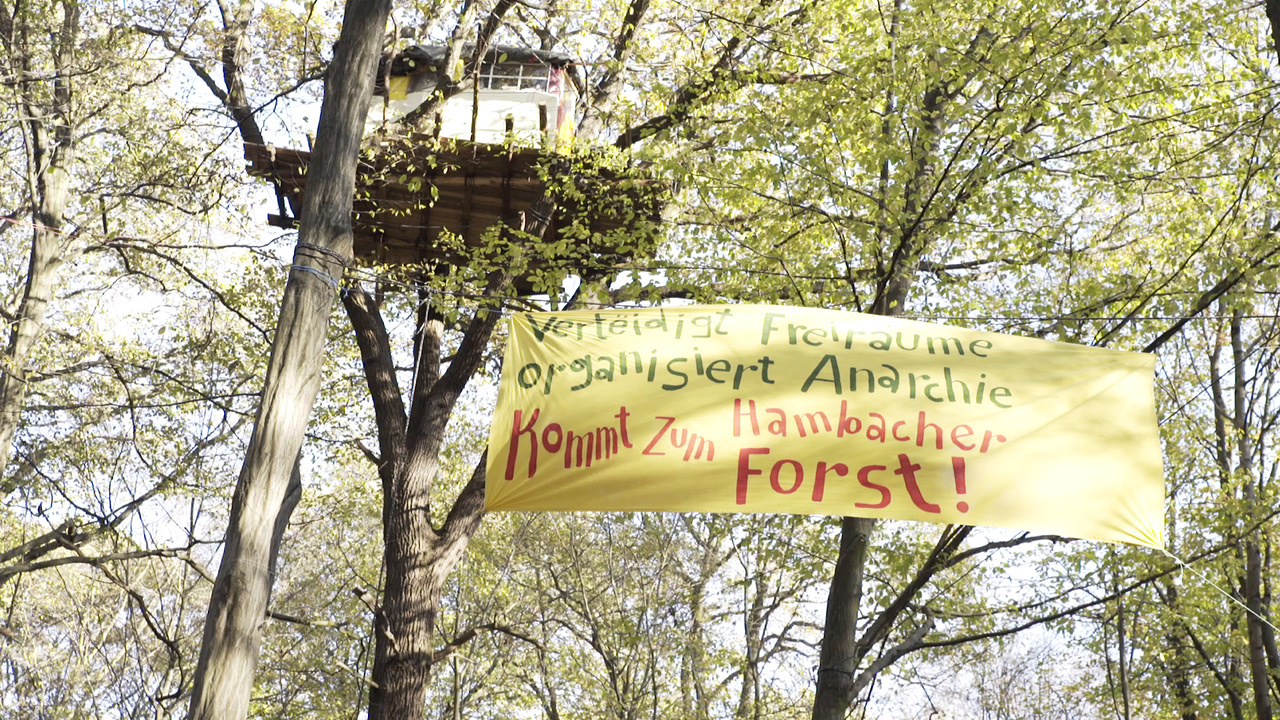 You can see a tree house. Below the tree house there is a banner stretched between the trees which reads: »Defend open spaces. Organizes anarchy. Come to the Hambach Forest!«