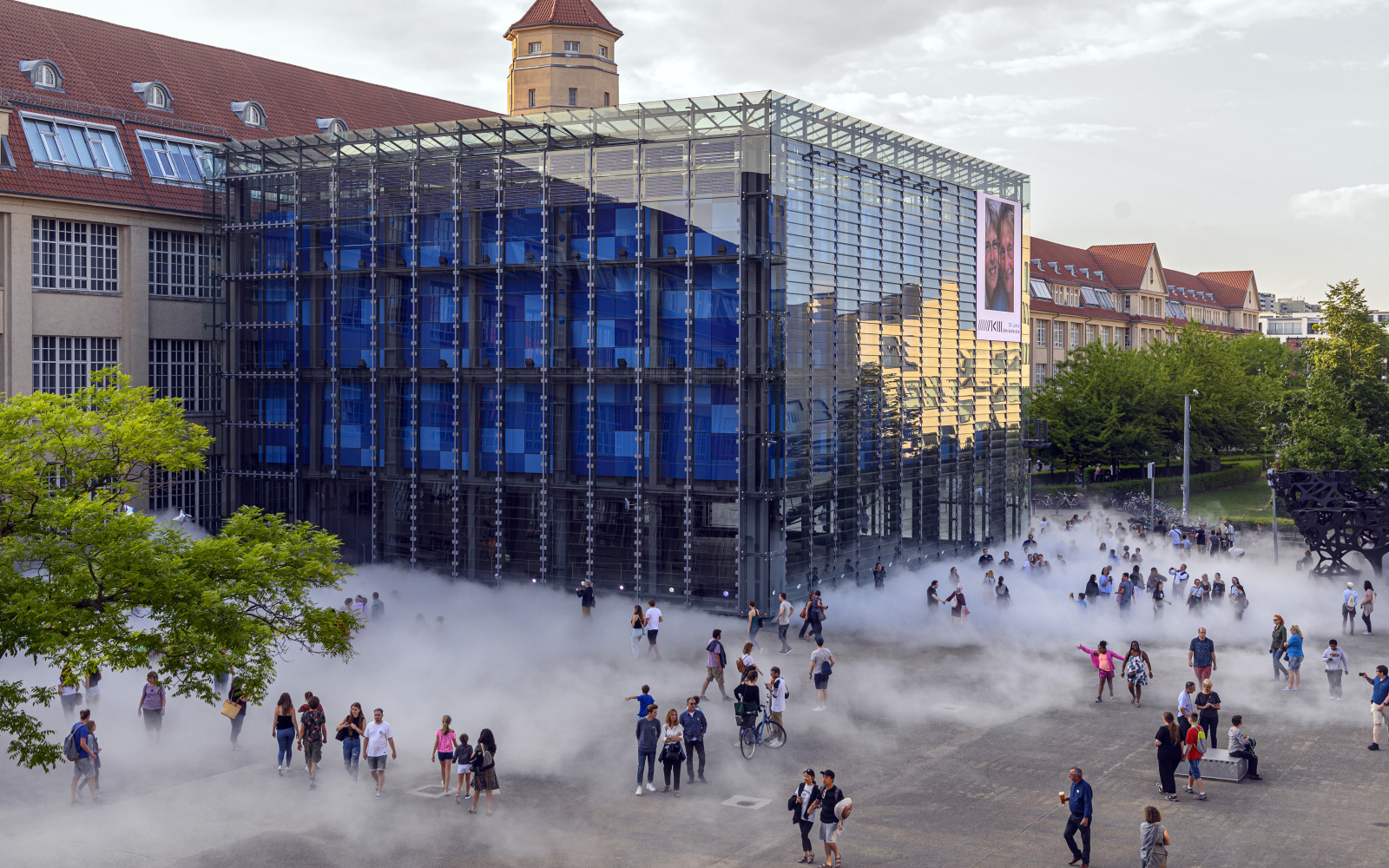 Das Foto wurde mit einer Drohne aufgenommen und zeigt den Kubus des ZKM bei Tag. Drum herum sind eine Menge Menschen versammelt die hinter der Nebelskulptur zum Teil verdeckt werden.