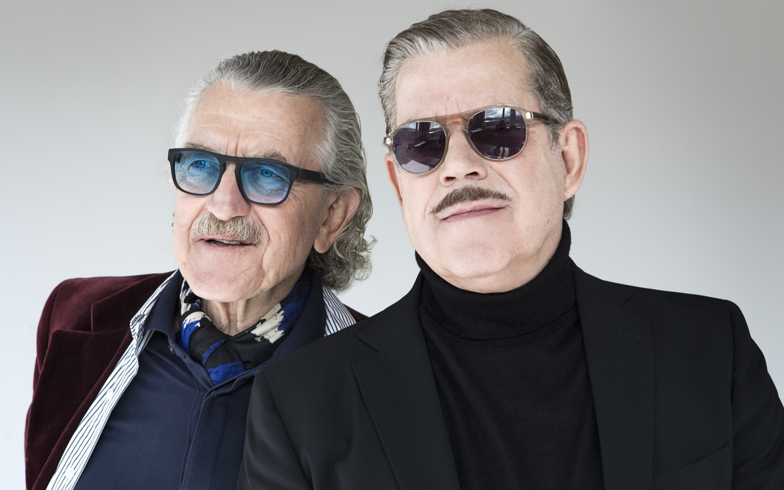 Zwei Männer stehen nebeneinander, die beiden Köpfe sind groß zu sehen. Der rechte trägt eine Sonnenbrille, einen Oberlippenbart, kurze, nach hinten gekämmte Haare und einen Rollkragen mit Jacket. Der linke trägt eine gefärbte Sonnenbrille.