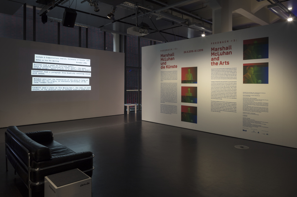 In the foreground on the left side is a black sofa. At the back left there is a partition wall with text projection. In the back on the right there is a wall printed with text and pictures.