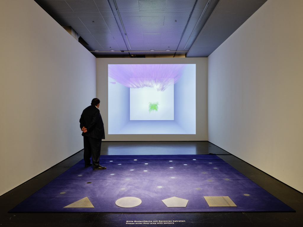 The photo shows Peter Weibel in front of his installation. A purple carpet with several dots on the floor can be seen. In the front there is a triangle, a circle, a pentagon and a square. A canvas shows a pink gradient and a square.