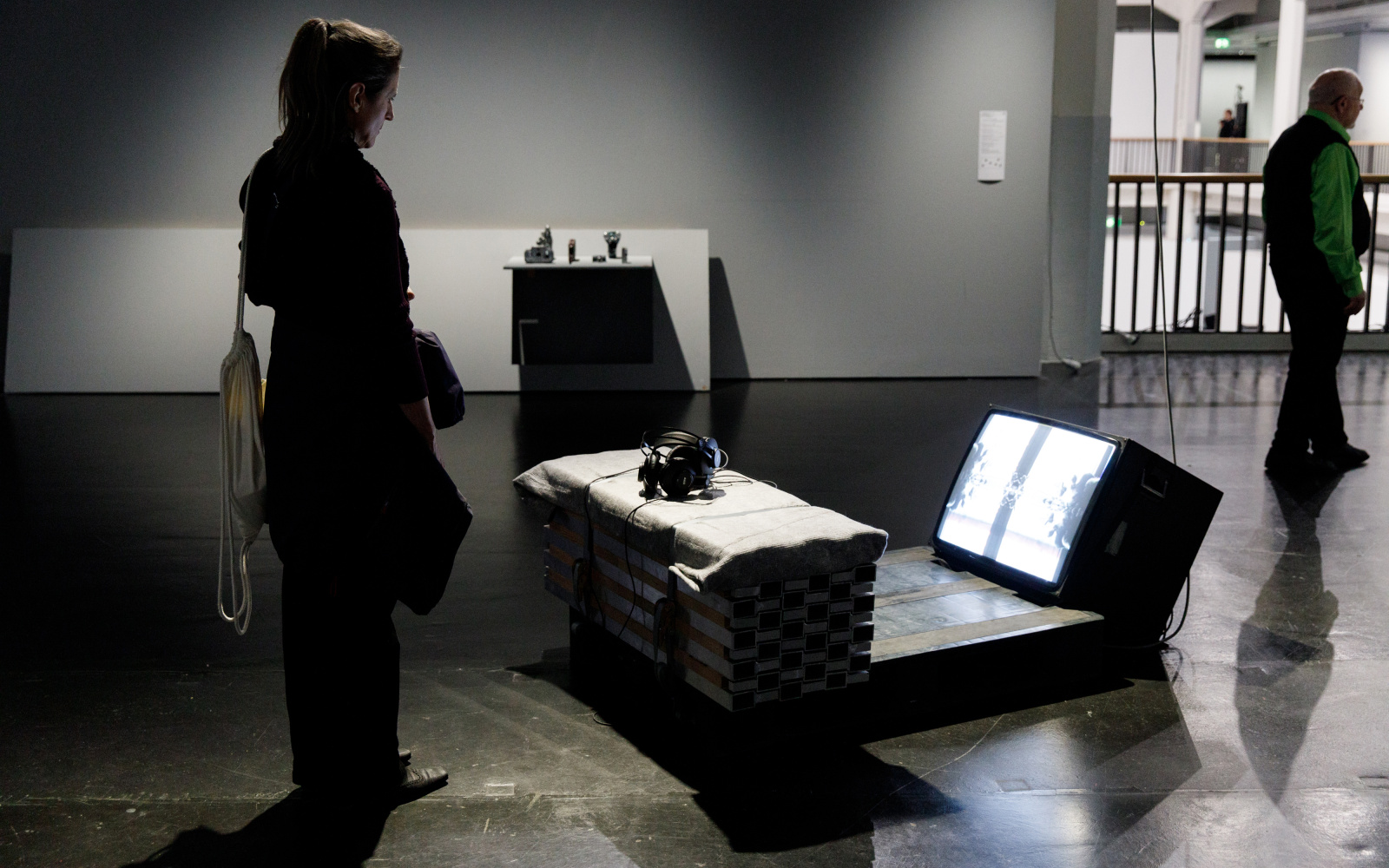A woman stands in front of an installation and looks at it. The installation consists of an improvised looking bench with headphones on it. In front of it there is a television set on the floor with its screen facing 45° upwards. 