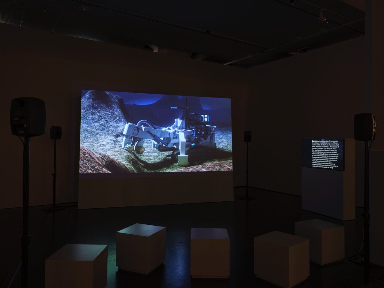 Das Bild zeigt einen Raum mit Sitzkästen, Monitoren und einem großen Bildschirm auf dem der Film »Prospecting Ocean« von Armin Linke abgespielt wird.