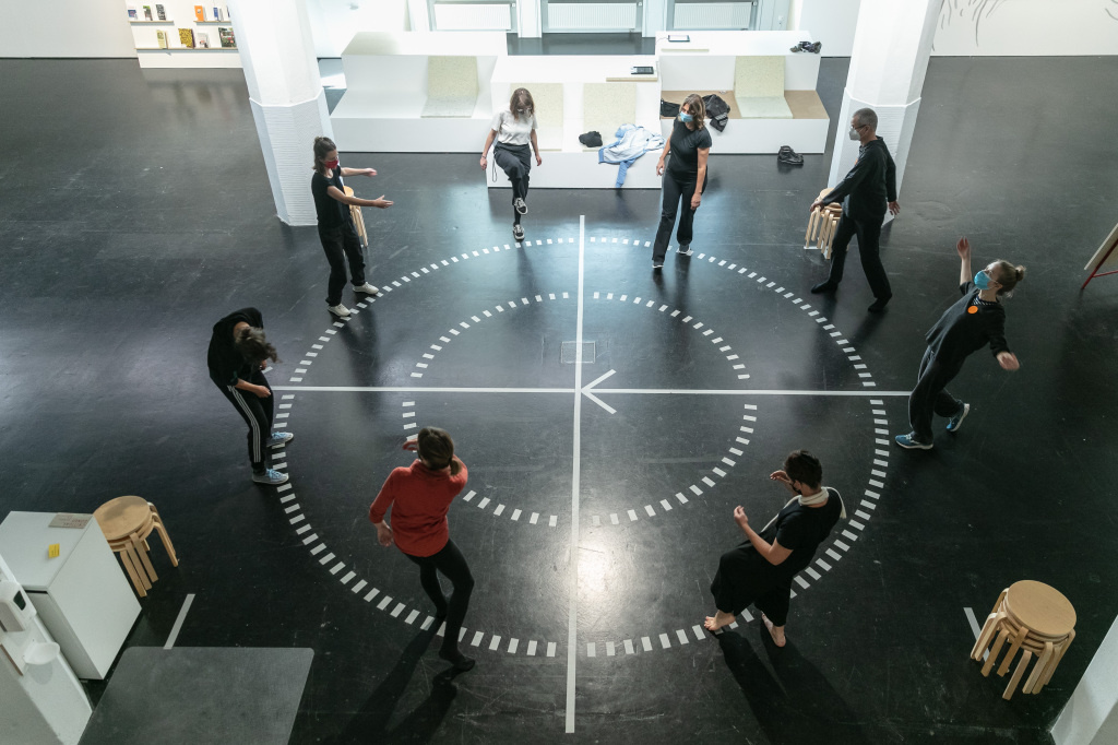 Eine Gruppe Menschen steht im Kreis auf einem runden Diagramm auf einem Hallenboden. Die Menschen scheinen in Bewegung zu sein.