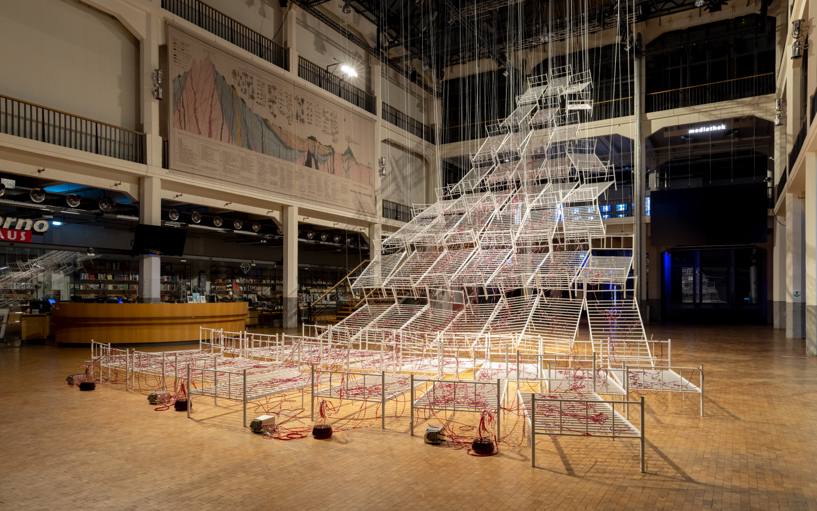 Installation view of Chiharu Shiota's »Connected to Life«. On view are several hospital bed frames hanging from the ceiling. Red liquid flows through transparent tubes on the beds.