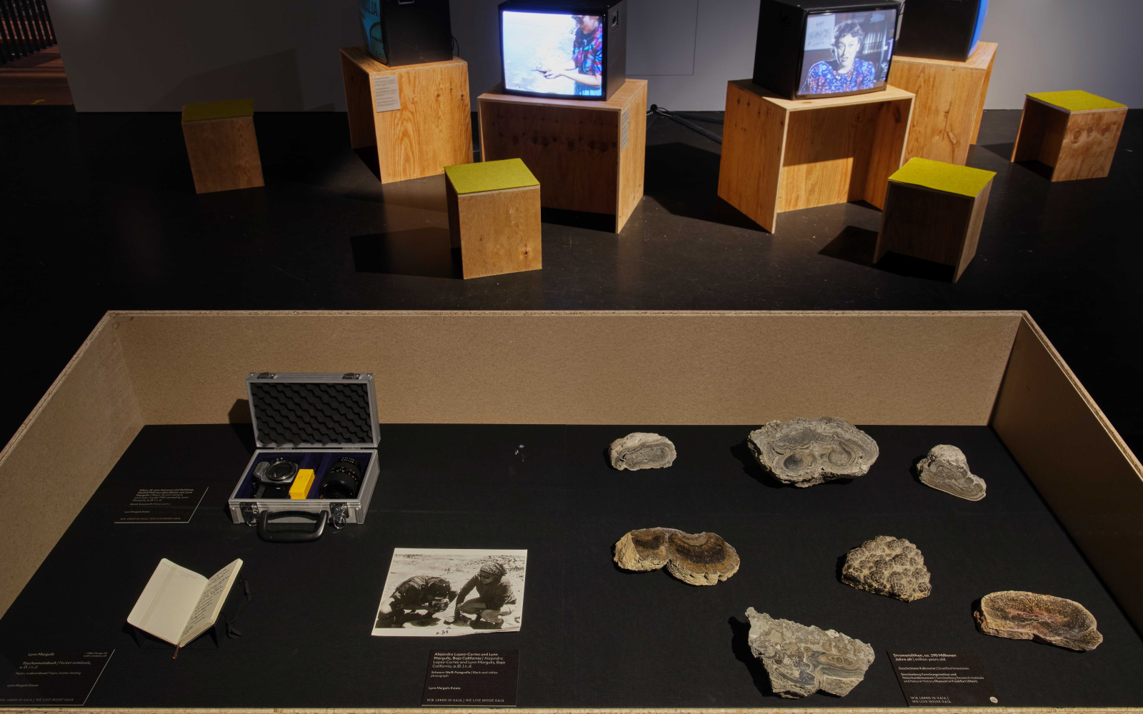 Foto aus der Ausstellung »Critical Zones« im Hintergrund laufen Videos auf Röhrenbildschirmen, im Vordergrund liegen Fotos und Gesteinsproben.