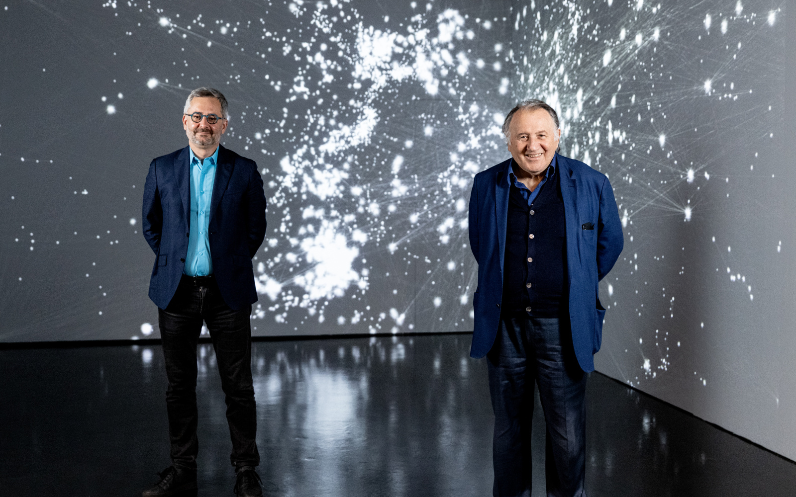 Zwei Männer stehen im Ausstellungsraum vor einer Projektion, die den Kosmos als Netzwerk zeigt. Der linke Mann ist der Wissenschaftler Albert-Lázsló Barabási. Der rechte Mann ist Peter Weibel. Beide lächeln, die Arme sind am Rücken.