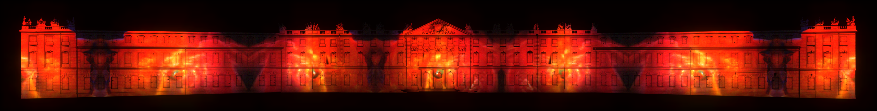Auf der Karlsruher Schlossfassade ist ein Projection Mapping zu sehen, das in der Nacht leuchtet. Das Mapping ist völlig Rot mit orangenen Elementen, eventuell wird Feuer dargestellt.