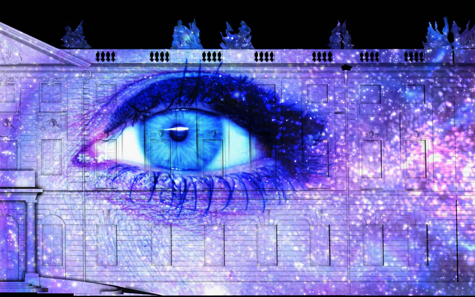 Auf der Karlsruher Schlossfassade ist ein Projection Mapping zu sehen, das in der Nacht leuchtet. Das Mapping zeigt das Gesicht einer Frau in der Mitte, die Augen sind weit geöffnet; um sie herum ist der Kosmos.